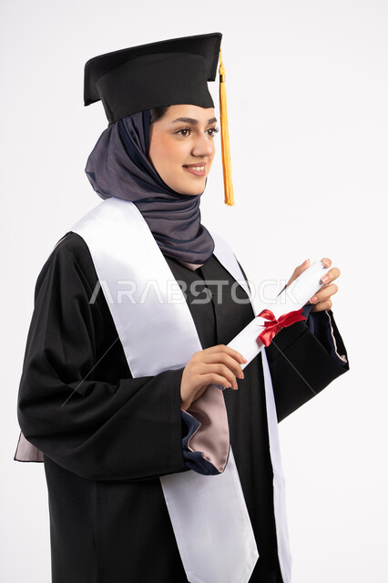 مفهوم النجاح والتفوق، الاحتفال بإنهاء الدراسة الجامعية، مناسبة واحتفال جامعي، شابة سعودية تنظر الى مكان ما بايماءات الفخر، تعابير السعادة بالوصول الى الحلم، بورتريه لخريجة عربية خليجية اماراتية محجبة ترتدي زي التخرج والقبعة تحمل بيدها الشهادة، خلفية بيضاء