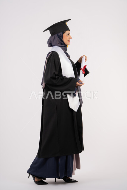الاحتفال بإنهاء الدراسة الجامعية، مناسبة واحتفال جامعي، شابة سعودية تنظر الى مكان ما بايماءات الفخر، تعابير السعادة بالوصول الى الحلم، مفهوم النجاح والتفوق، بورتريه من الجانب لخريجة عربية خليجية اماراتية محجبة ترتدي زي التخرج والقبعة تحمل بيدها الشهادة، خلفية بيضاء