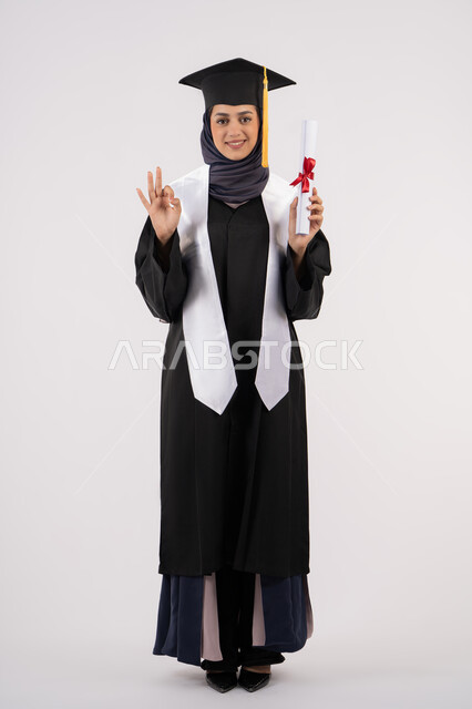 Mark of excellence and distinction, a young Saudi woman standing with gestures of pride and happiness for achieving her dream, the concept of success and excellence, celebrating the completion of university studies, a portrait of a veiled Arab Gulf Emirati graduate wearing a graduation gown and cap, holding a certificate in her hand, full body, white background