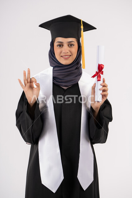 علامة الامتياز والتميز، تعابير السعادة بالوصول الى الحلم، شابة سعودية تنظر الى الكاميرا بايماءات الفخر، مفهوم النجاح والتفوق، الاحتفال بإنهاء الدراسة الجامعية، بورتريه لخريجة عربية خليجية اماراتية محجبة ترتدي زي التخرج والقبعة تحمل بيدها الشهادة، خلفية بيضاء