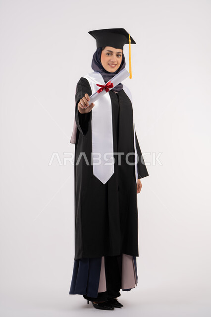 مناسبة واحتفال جامعي، شابة سعودية تنظر الى الكاميرا بايماءات الفخر، تعابير السعادة بالوصول الى الحلم، مفهوم النجاح والتفوق، الاحتفال بإنهاء الدراسة الجامعية، بورتريه لخريجة عربية خليجية اماراتية محجبة ترتدي زي التخرج والقبعة تحمل بيدها الشهادة، خلفية بيضاء