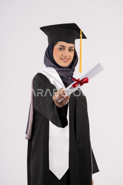 تعابير السعادة بالوصول الى الحلم، شابة سعودية تنظر الى الكاميرا بايماءات الفخر، مفهوم النجاح والتفوق، مناسبة واحتفال جامعي، الاحتفال بإنهاء الدراسة الجامعية، بورتريه لخريجة عربية خليجية اماراتية محجبة ترتدي زي التخرج والقبعة تحمل بيدها الشهادة، خلفية بيضاء
