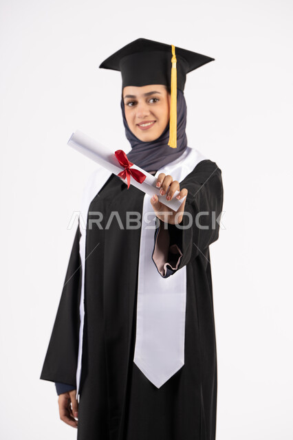 تعابير السعادة بالوصول الى الحلم، شابة سعودية تنظر الى الكاميرا بايماءات الفخر، مفهوم النجاح والتفوق، مناسبة واحتفال جامعي، الاحتفال بإنهاء الدراسة الجامعية، بورتريه لخريجة عربية خليجية اماراتية محجبة ترتدي زي التخرج والقبعة تحمل بيدها الشهادة، خلفية بيضاء