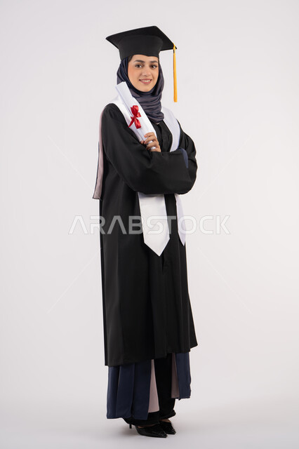 الاحتفال بإنهاء الدراسة الجامعية، إيماءات السعادة بالوصول الى الحلم، مناسبة واحتفال جامعي، مفهوم النجاح والتفوق، شابة سعودية تقف بأيدي متقاطعة بتعابير الفخر، بورتريه لخريجة عربية خليجية اماراتية محجبة ترتدي زي التخرج والقبعة تحمل بيدها الشهادة، خلفية بيضاء