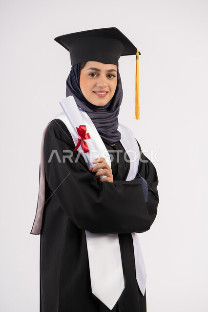 إيماءات السعادة بالوصول الى الحلم، مناسبة واحتفال جامعي، مفهوم النجاح والتفوق، شابة سعودية تقف بأيدي متقاطعة بتعابير الفخر، الاحتفال بإنهاء الدراسة الجامعية، بورتريه لخريجة عربية خليجية اماراتية محجبة ترتدي زي التخرج والقبعة تحمل بيدها الشهادة، خلفية بيضاء