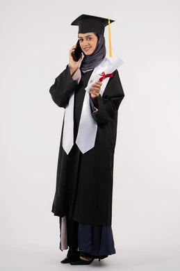 Sharing the joy of achieving the dream of success with family and friends, a young Saudi woman using a modern technical device, a portrait of a veiled Arab Gulf Emirati graduate wearing a graduation gown and a hat, holding a certificate in her hand, making a phone call via mobile phone, full body, white background