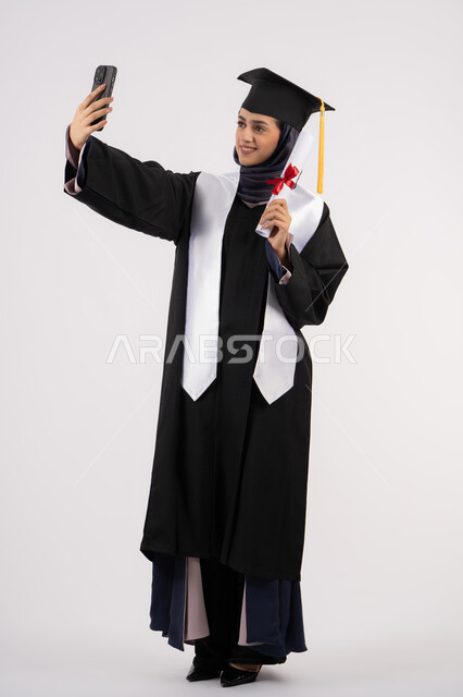 التفوق والنجاح والاجتهاد، توثيق لحظة النجاح عبر الهاتف المحمول، خريجة اماراتية تمسك الشهادة تحتفل بإنهاء المرحلة الجامعية، بورتريه لشابة عربية خليجية سعودية مبتسمة ترتدي القبعة وعباءة التخرج تقوم بالتقاط صور سيلفي بتعابير السرور، الجسم بالطول الكامل، خلفية بيضاء