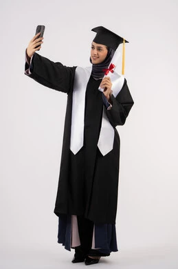 التفوق والنجاح والاجتهاد، توثيق لحظة النجاح عبر الهاتف المحمول، خريجة اماراتية تمسك الشهادة تحتفل بإنهاء المرحلة الجامعية، بورتريه لشابة عربية خليجية سعودية مبتسمة ترتدي القبعة وعباءة التخرج تقوم بالتقاط صور سيلفي بتعابير السرور، الجسم بالطول الكامل، خلفية بيضاء