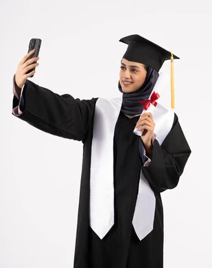 توثيق لحظة النجاح عبر الهاتف المحمول، خريجة اماراتية تمسك الشهادة تحتفل بإنهاء المرحلة الجامعية، بورتريه لشابة عربية خليجية سعودية مبتسمة ترتدي القبعة وعباءة التخرج تقوم بالتقاط صور سيلفي بتعابير السرور، التفوق والنجاح والاجتهاد، خلفية بيضاء