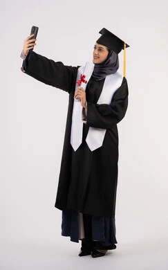 Excellence, success and hard work, documenting the moment of success via mobile phone, Emirati graduate holding her certificate celebrating the completion of university, portrait of a smiling young Saudi Arabian Gulf Arab woman wearing a graduation cap and gown taking selfies with happy expressions, full body, white background