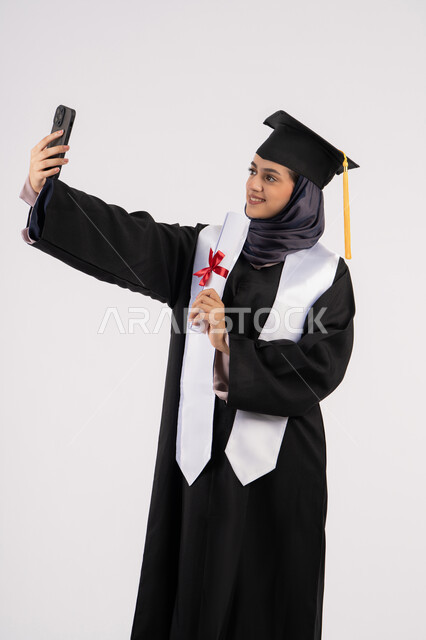 توثيق لحظة النجاح عبر الهاتف المحمول، خريجة اماراتية تمسك الشهادة تحتفل بإنهاء المرحلة الجامعية، بورتريه لشابة عربية خليجية سعودية مبتسمة ترتدي القبعة وعباءة التخرج تقوم بالتقاط صور سيلفي بتعابير السرور، التفوق والنجاح والاجتهاد، خلفية بيضاء