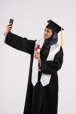 Documenting a moment of success via mobile phone, an Emirati graduate holding her diploma celebrating the completion of university, a portrait of a smiling young Saudi Arabian Gulf Arab woman wearing a graduation cap and gown taking a selfie with expressions of joy, excellence, success and diligence,, white background