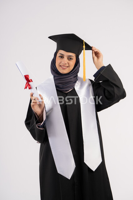 شابة سعودية ننظر الى الكاميرا بايماءات الفخر، مفهوم النجاح والتفوق، إيماءات السعادة بالوصول الى الحلم، مناسبة واحتفال جامعي، الاحتفال بإنهاء الدراسة الجامعية، بورتريه لخريجة عربية خليجية اماراتية محجبة ترتدي زي التخرج والقبعة تحمل بيدها الشهادة، خلفية بيضاء