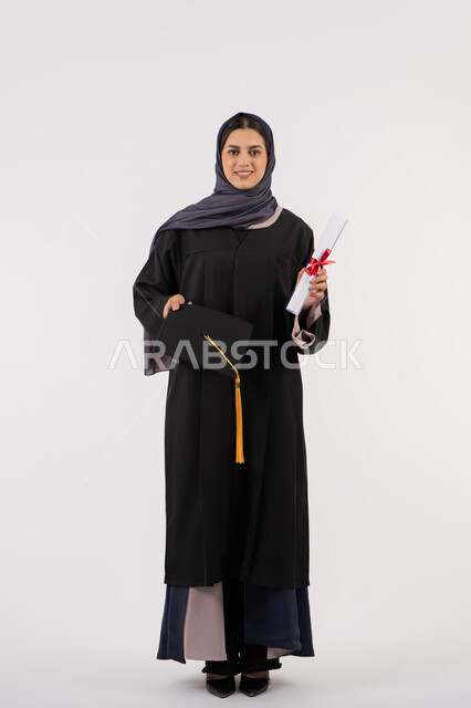الاستعداد لرمي القبعة احتفالا بالنجاح وانتهاء المرحلة الجامعية، بورتريه لخريجة عربية خليجية اماراتية ترتدي قبعة وزي التخرج تمسك بيدها الشهادة، شابة سعودية تقف بتعابير وايماءات السرور والسعادة، مفهوم التفوق والاجتهاد، الجسم بالطول الكامل، خلفية بيضاء