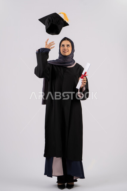 رمي القبعة احتفالا بالنجاح وانتهاء المرحلة الجامعية، بورتريه لخريجة عربية خليجية اماراتية ترتدي قبعة وزي التخرج تمسك بيدها الشهادة، شابة سعودية تقف بتعابير وايماءات السرور والسعادة، مفهوم التفوق والاجتهاد، الجسم بالطول الكامل، خلفية بيضاء