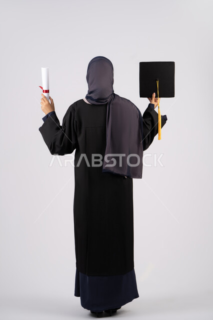انتهاء مرحلة الدراسة الجامعية، الوصول للحلم والنجاح، التفوق والاجتهاد والجد، بورتريه مقرب من الخلف لشابة عربية خليجية سعودية محجبة ترتدي زي التخرج تمسك بيدها الشهادة والقبعة، خريجة اماراتية تحتفل بتخرجها من الجامعة، الجسم بالطول الكامل، خلفية بيضاء