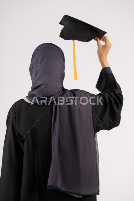 الوصول للحلم والنجاح، انتهاء مرحلة الدراسة الجامعية، التفوق والاجتهاد والجد، بورتريه مقرب من الخلف لشابة عربية خليجية سعودية محجبة ترتدي زي التخرج تمسك بيدها الشهادة والقبعة، خريجة اماراتية تحتفل بتخرجها من الجامعة، خلفية بيضاء