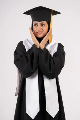 Looking at a place, gestures and expressions of pleasure and happiness, portrait of a smiling young Saudi Arabian Gulf woman wearing a graduation cap and gown, placing her hands under her face with feminine gestures, celebrating university graduation, an Emirati graduate finishing her university studies, white background