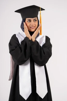 النظر الى مكان ما، حركات وتعابير السرور والسعادة، بورتريه لشابة عربية خليجية سعودية مبتسمة ترتدي قبعة وزي التخرج تضع يديها اسفل وجهها بايماءات الانوثة، الاحتفال بالتخرج من الجامعة، خريجة اماراتية تنهي مرحلة الدراسة الجامعية، خلفية بيضاء