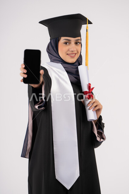 Blank black screen on mobile phone, portrait of a young Saudi Arabian Gulf Arab woman wearing a cap and graduation gown holding a diploma, smiling Emirati graduate standing with an expression of pleasure and happiness, online study applications, celebrating the end of the university stage, white background