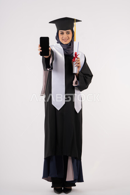 Online study applications, displaying a blank black screen on a mobile phone, portrait of a young Saudi Arabian Gulf Arab woman wearing a cap and graduation gown holding a diploma, a smiling Emirati graduate standing with an expression of pleasure and happiness, celebrating the end of the university stage, full body, white background