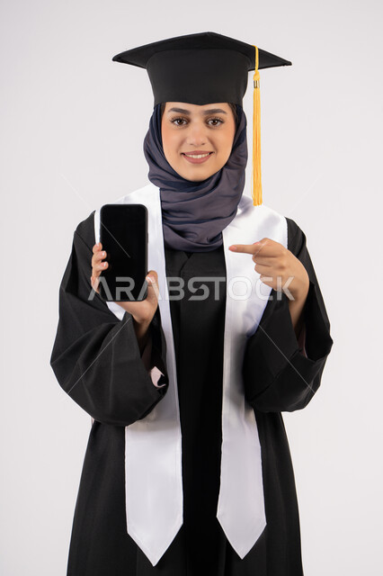 الاشارة الى شاشة سوداء فارغة على الهاتف المحمول، بورتريه لشابة عربية خليجية سعودية ترتدي القبعة وزي التخرج تنظر الى الكاميرا، خريجة اماراتية مبتسمة تقف بتعابير السرور والسعادة، تطبيقات الدراسة اونلاين، الاحتفال بانتهاء المرحلة الجامعية، خلفية بيضاء