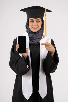 الاشارة الى شاشة سوداء فارغة على الهاتف المحمول، بورتريه لشابة عربية خليجية سعودية ترتدي القبعة وزي التخرج تنظر الى الكاميرا، خريجة اماراتية مبتسمة تقف بتعابير السرور والسعادة، تطبيقات الدراسة اونلاين، الاحتفال بانتهاء المرحلة الجامعية، خلفية بيضاء