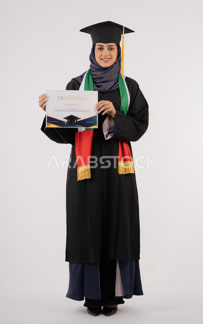 النظر الى الكاميرا بايماءات الفخر، النجاح والتفوق والتخرج من الجامعة، إيماءات السعادة بالوصول الى الحلم، مناسبة واحتفال جامعي، الاحتفال بإنهاء الدراسة الجامعية، بورتريه لخريجة عربية خليجية اماراتية محجبة ترتدي زي التخرج والقبعة تحمل بيدها الشهادة، خلفية بيضاء