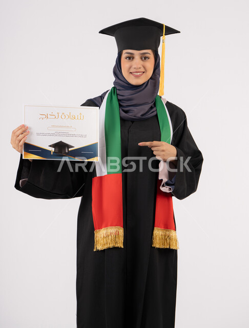 الاشارة باصبع السبابة إلى الشهادة، النجاح والتفوق والتخرج من الجامعة، إيماءات السعادة بالوصول الى الحلم، مناسبة واحتفال جامعي، الاحتفال بإنهاء الدراسة الجامعية، بورتريه لخريجة عربية خليجية اماراتية محجبة ترتدي زي التخرج والقبعة تنظر الى الكاميرا، خلفية بيضاء