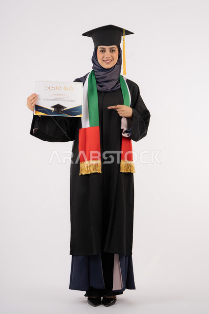 الاشارة باصبع السبابة إلى الشهادة، النجاح والتفوق والتخرج من الجامعة، إيماءات السعادة بالوصول الى الحلم، مناسبة واحتفال جامعي، الاحتفال بإنهاء الدراسة الجامعية، بورتريه لخريجة عربية خليجية اماراتية محجبة ترتدي زي التخرج والقبعة تنظر الى الكاميرا، خلفية بيضاء