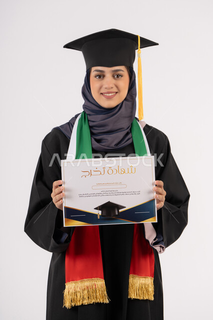 النظر الى الكاميرا بايماءات الفخر، النجاح والتفوق والتخرج من الجامعة، إيماءات السعادة بالوصول الى الحلم، مناسبة واحتفال جامعي، الاحتفال بإنهاء الدراسة الجامعية، بورتريه لخريجة عربية خليجية اماراتية محجبة ترتدي زي التخرج والقبعة تحمل بيدها الشهادة، خلفية بيضاء