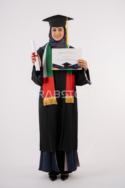 Success, excellence, and graduation from university, gestures of happiness for achieving a dream, a university occasion and celebration, celebrating the completion of university studies, looking at the camera with gestures of pride, a portrait of a veiled Arab Gulf Emirati graduate wearing a graduation gown and cap, holding a certificate in her hand, white background
