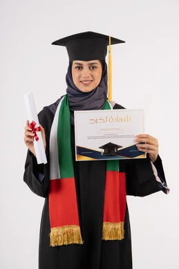 Looking at the camera with gestures of pride, success, excellence and graduation from university, gestures of happiness for achieving the dream, university occasion and celebration, celebration of completing university studies, portrait of a veiled Arab Gulf Emirati female graduate wearing graduation attire and cap holding the certificate in her hand, white background