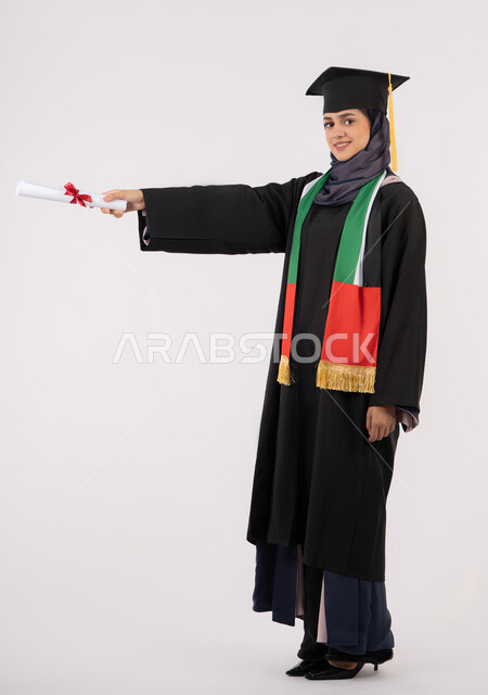 الشعور بالفخر والثقة بالنفس، إيماءات السعادة بالوصول لحلم النجاح، مناسبة واحتفال جامعي، الاحتفال بإنهاء الدراسة الجامعية، مفهوم الاجتهاد والتفوق، بورتريه لخريجة عربية خليجية اماراتية محجبة ترتدي زي التخرج والقبعة تمسك بيدها شهادة التخرج، خلفية بيضاء
