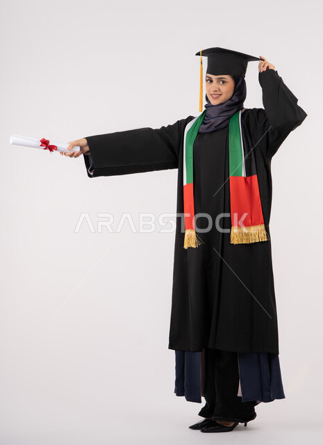النجاح والتفوق والتخرج من الجامعة، إيماءات السعادة بالوصول الى الحلم، مناسبة واحتفال جامعي، الاحتفال بإنهاء الدراسة الجامعية، النظر الى الكاميرا بايماءات الفخر، بورتريه لخريجة عربية خليجية اماراتية محجبة ترتدي زي التخرج والقبعة تحمل بيدها الشهادة، خلفية بيضاء