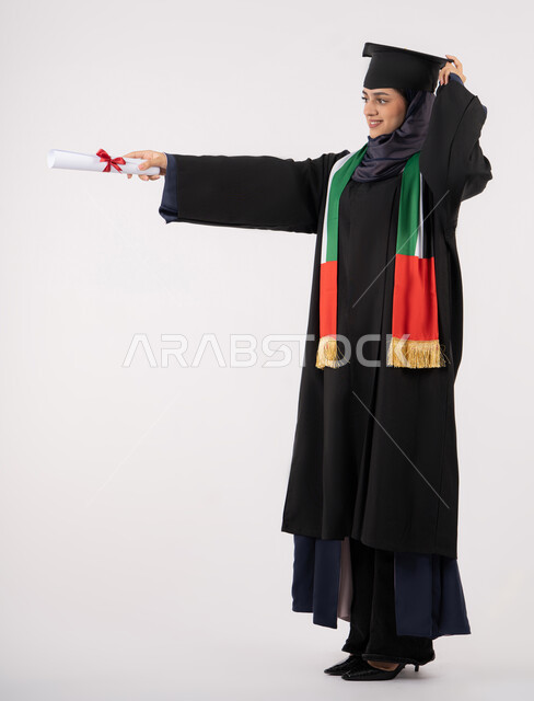 النظر الى مكان ما بايماءات الفخر، النجاح والتفوق والتخرج من الجامعة، إيماءات السعادة بالوصول الى الحلم، مناسبة واحتفال جامعي، الاحتفال بإنهاء الدراسة الجامعية، بورتريه لخريجة عربية خليجية اماراتية محجبة ترتدي زي التخرج والقبعة تحمل بيدها الشهادة، خلفية بيضاء