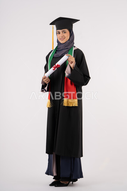 Success, excellence, and graduation from university, gestures of happiness for achieving a dream, a university occasion and celebration, celebrating the completion of university studies, looking at the camera with gestures of pride, a portrait of a veiled Arab Gulf Emirati graduate wearing a graduation gown and cap, holding a certificate in her hand, white background