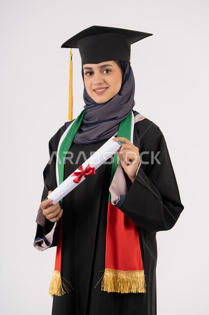 الشعور بالفخر والثقة بالنفس، إيماءات السعادة بالوصول لحلم النجاح، مناسبة واحتفال جامعي، الاحتفال بإنهاء الدراسة الجامعية، مفهوم الاجتهاد والتفوق، بورتريه لخريجة عربية خليجية اماراتية محجبة ترتدي زي التخرج والقبعة تمسك بيدها شهادة التخرج، خلفية بيضاء