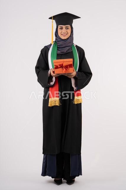 تعابير السعادة والسرور بالوصول لحلم النجاح، تقديم الهدايا في المناسبات، مفهوم الاجتهاد والتفوق، الاحتفال بإنهاء الدراسة الجامعية، مناسبة واحتفال جامعي، بورتريه لخريجة عربية خليجية اماراتية محجبة ترتدي زي التخرج والقبعة تمسك بيدها صندوق الهدية، خلفية بيضاء
