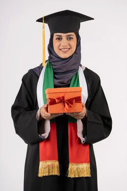 Giving gifts on occasions, the concept of hard work and excellence, expressions of happiness and joy at achieving the dream of success, celebrating the completion of university studies, a university occasion and celebration, a portrait of a veiled Arab Gulf Emirati graduate wearing a graduation gown and hat, holding a gift box in her hand, white background