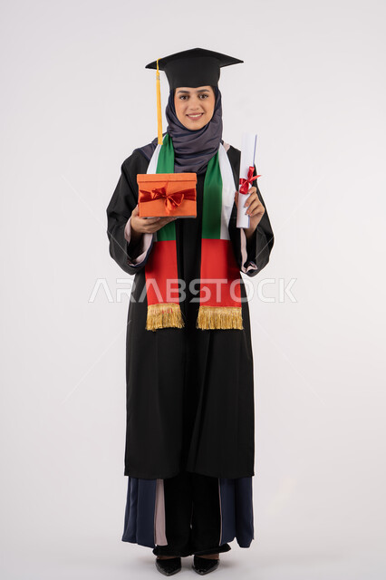 الاحتفال بإنهاء الدراسة الجامعية، مفهوم الاجتهاد والتفوق، تعابير السعادة بالوصول لحلم النجاح، تقديم الهدايا في المناسبات، مناسبة واحتفال جامعي، بورتريه لخريجة عربية خليجية اماراتية محجبة ترتدي زي التخرج والقبعة تمسك بيدها صندوق الهدية مع الشهادة، خلفية بيضاء