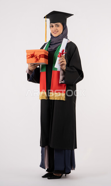 الوصول لحلم النجاح، الاحتفال بإنهاء الدراسة الجامعية، مفهوم الاجتهاد والتفوق، تقديم الهدايا في المناسبات، مناسبة واحتفال جامعي، بورتريه لخريجة عربية خليجية اماراتية محجبة ترتدي زي التخرج والقبعة تمسك بيدها صندوق الهدية مع الشهادة، خلفية بيضاء