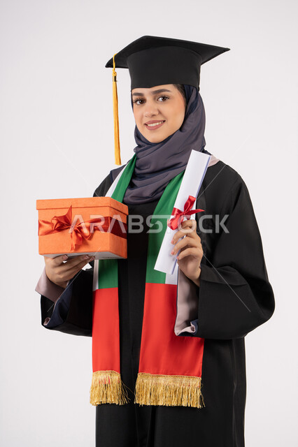 The concept of diligence and excellence, expressions of happiness at achieving the dream of success, celebrating the completion of university studies, giving gifts on occasions, a university occasion and celebration, a portrait of a veiled Arab Gulf Emirati graduate wearing a graduation gown and cap, holding a gift box with a certificate, white background