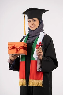 The concept of diligence and excellence, expressions of happiness at achieving the dream of success, celebrating the completion of university studies, giving gifts on occasions, a university occasion and celebration, a portrait of a veiled Arab Gulf Emirati graduate wearing a graduation gown and cap, holding a gift box with a certificate, white background
