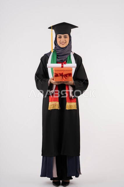 الاحتفال بإنهاء الدراسة الجامعية، مفهوم الاجتهاد والتفوق، تعابير السعادة بالوصول لحلم النجاح، تقديم الهدايا في المناسبات، مناسبة واحتفال جامعي، بورتريه لخريجة عربية خليجية اماراتية محجبة ترتدي زي التخرج والقبعة تمسك بيدها صندوق الهدية مع الشهادة، خلفية بيضاء