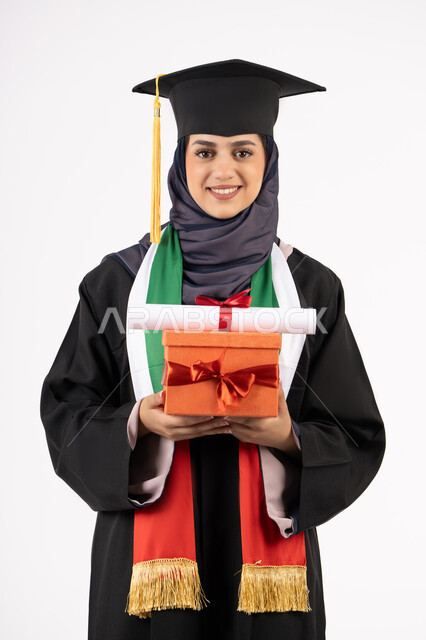 مفهوم الاجتهاد والتفوق، تعابير السعادة بالوصول لحلم النجاح، الاحتفال بإنهاء الدراسة الجامعية، تقديم الهدايا في المناسبات، مناسبة واحتفال جامعي، بورتريه لخريجة عربية خليجية اماراتية محجبة ترتدي زي التخرج والقبعة تمسك بيدها صندوق الهدية مع الشهادة، خلفية بيضاء