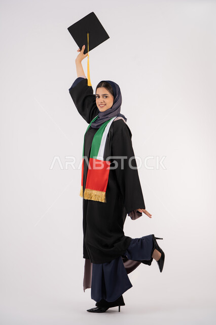 المناسبات والاحتفالات الجامعية، النظر الى الكاميرا بايماءات الفخر، الوصول إلى حلم النجاح، مفهوم الاجتهاد والتفوق، الاحتفال بإنهاء الدراسة الجامعية، مناسبة واحتفال جامعي، بورتريه لخريجة عربية خليجية اماراتية محجبة ترتدي زي التخرج تمسك بيدها القبعة، خلفية بيضاء