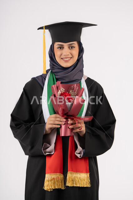 الاحتفال بإنهاء الدراسة الجامعية، إيماءات السعادة بالوصول لحلم النجاح، بورتريه لخريجة عربية خليجية اماراتية محجبة ترتدي زي التخرج والقبعة تمسك بيدها بوكيه ورد تنظر إلى الكاميرا بتعابير السرور، مناسبة واحتفال جامعي، خلفية بيضاء