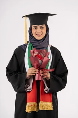 Celebrating the completion of university studies, gestures of happiness for reaching the dream of success, portrait of a veiled Arab Gulf Emirati graduate wearing a graduation gown and cap, holding a bouquet of flowers in her hand, looking at the camera with an expression of pleasure, a university occasion and celebration, white background