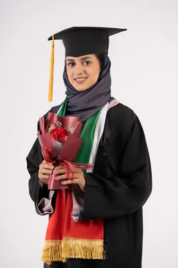 إيماءات السعادة بالوصول لحلم النجاح، بورتريه لخريجة عربية خليجية اماراتية محجبة ترتدي زي التخرج والقبعة تمسك بيدها بوكيه ورد تنظر إلى الكاميرا بتعابير السرور، الاحتفال بإنهاء الدراسة الجامعية، مناسبة واحتفال جامعي، خلفية بيضاء